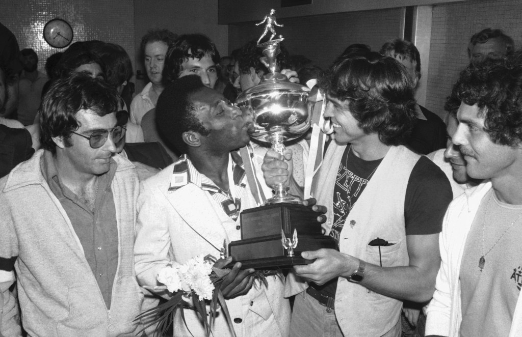 Black and white photo of Pelé kissing a championship trophy while celebrating with New York Cosmos teammates and staff. The group is surrounded by jubilant fans in a crowded room, capturing a historic moment of victory and legacy in American soccer.
